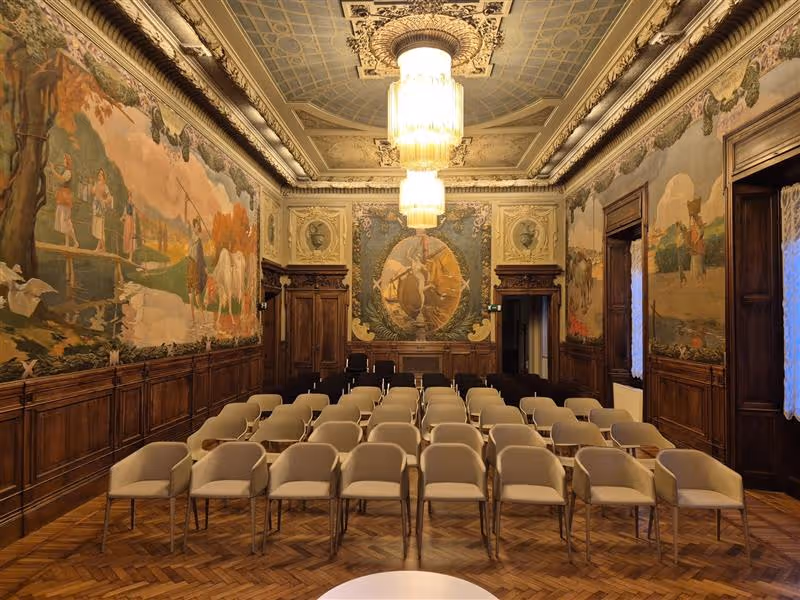 Sala conferenze storica con pareti decorate da affreschi e sedie beige disposte in file su pavimento in parquet.