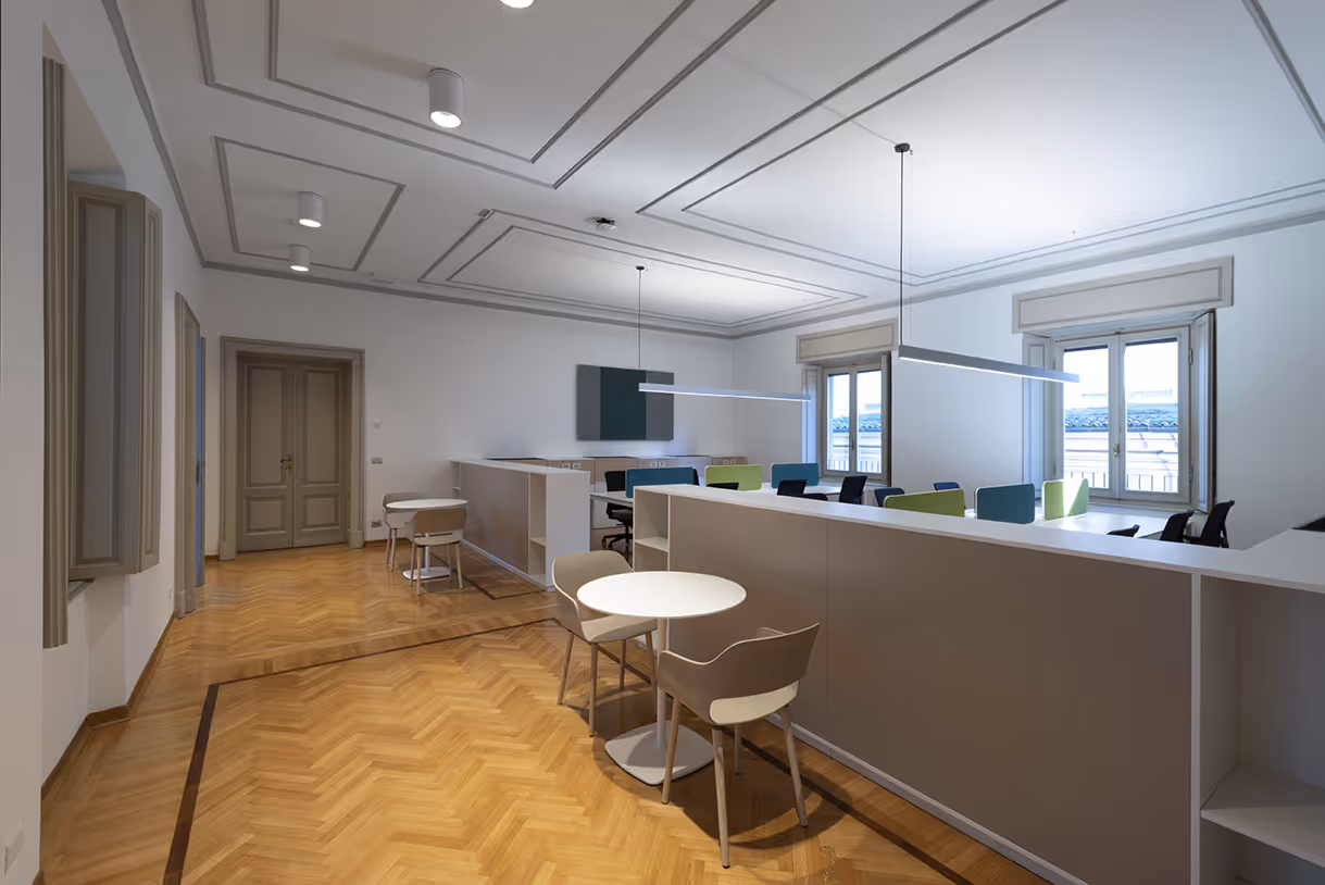 Ufficio moderno con pavimento in legno a spina di pesce, scrivanie bianche, sedie di vari colori e grandi finestre che lasciano entrare luce naturale.