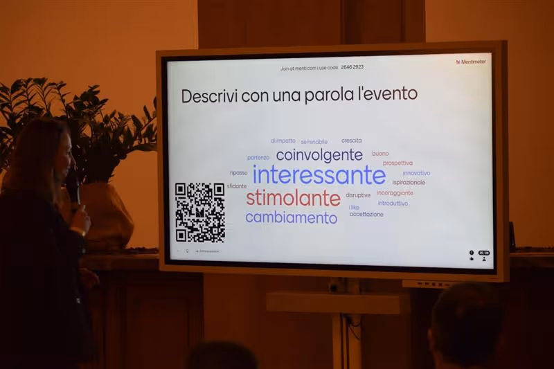 Uomo che presenta con microfono davanti a un pubblico seduto in una sala conferenze moderna con schermo digitale sullo sfondo.