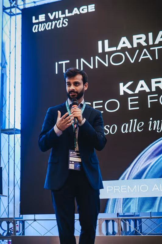 Uomo che presenta con microfono davanti a un pubblico seduto in una sala conferenze moderna con schermo digitale sullo sfondo.