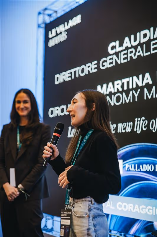 Donna che parla con un microfono in mano durante una conferenza, con un'altra donna sorridente sullo sfondo e un grande schermo con testo.