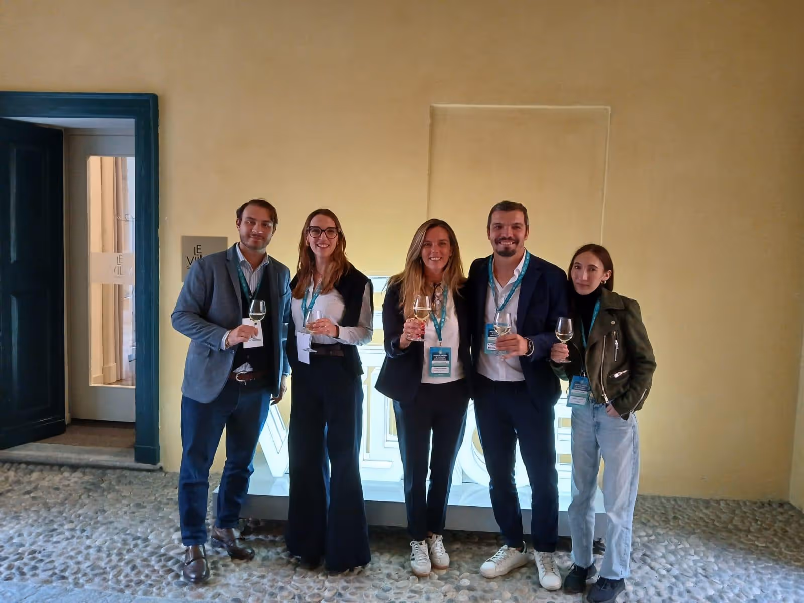 Cinque persone in piedi davanti a un'insegna luminosa con la scritta Welcome, con calici in mano, sorridenti in un ambiente interno.