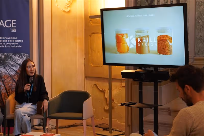 Uomo che presenta con microfono davanti a un pubblico seduto in una sala conferenze moderna con schermo digitale sullo sfondo.