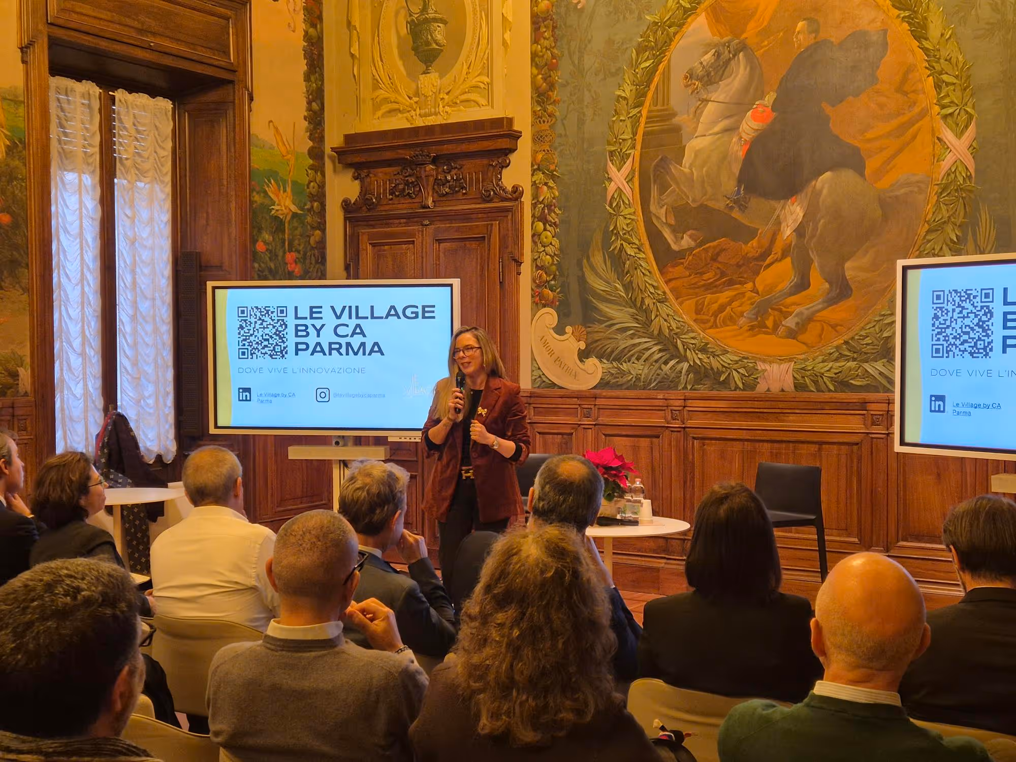Donna che presenta con microfono davanti a un pubblico in una sala storica decorata con un grande dipinto e due schermi con il logo di Le Village by CA Parma.