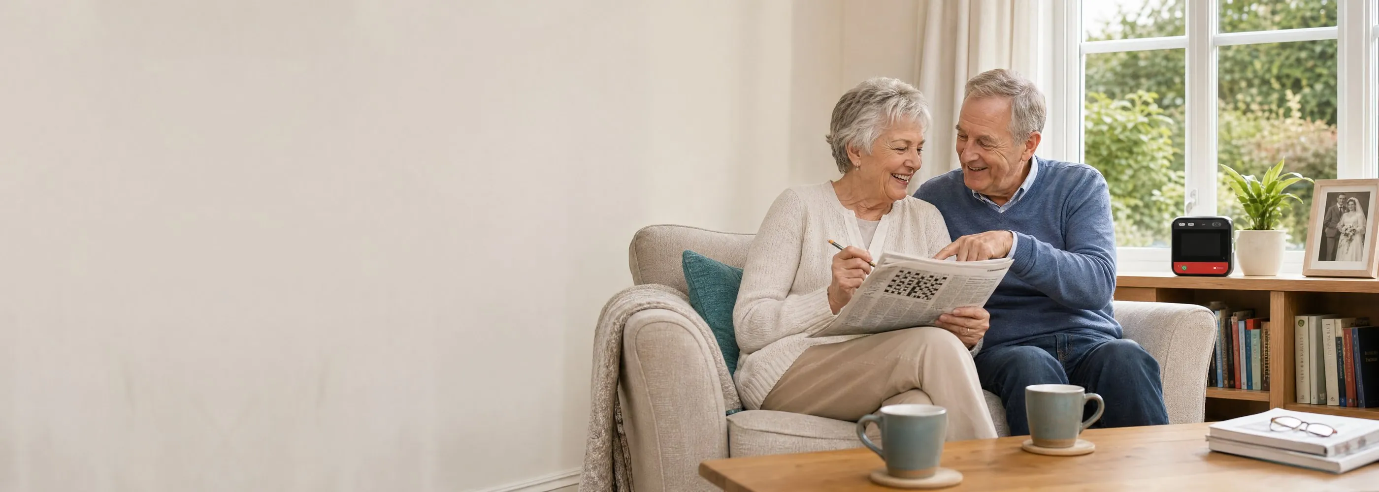 An older couple doing the crossword on the sofa, with their home defibrillator on the bookcase beside the window