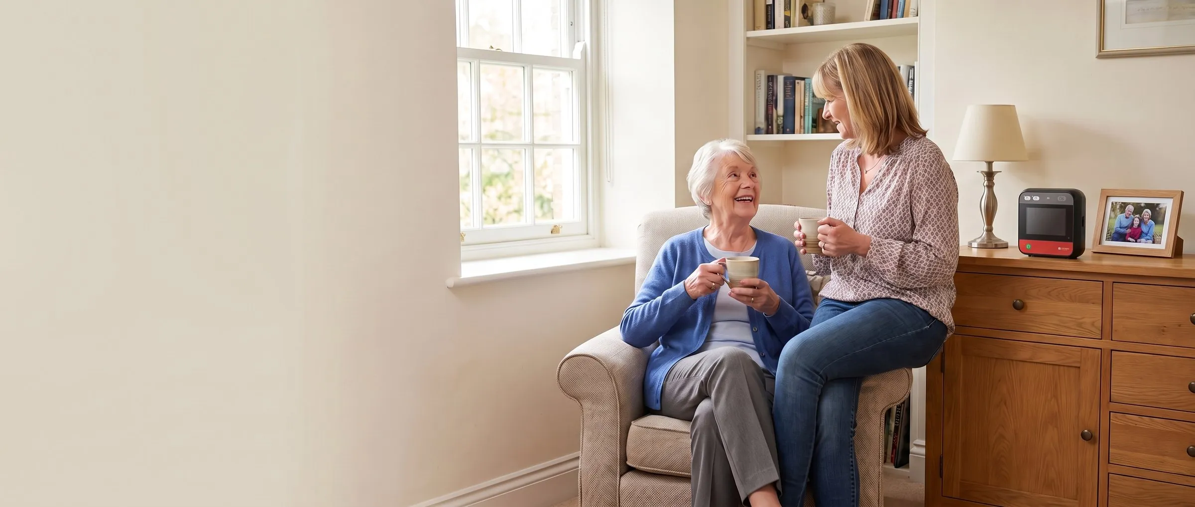 A mother and her grown-up daughter chatting over tea at home, with a home defibrillator on the sideboard behind them
