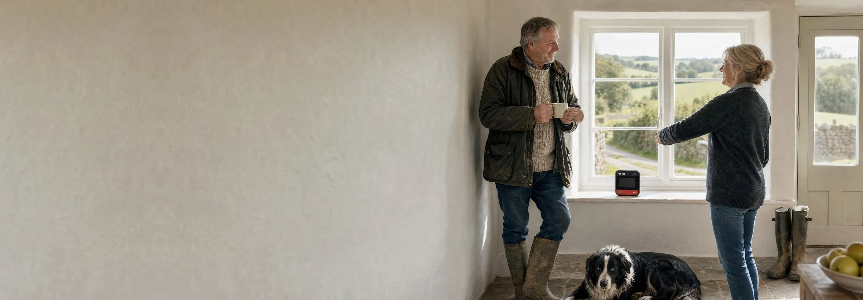 A farming couple chatting over a mug of tea in their farmhouse kitchen, with their sheepdog on the floor and their home defibrillator on the windowsill overlooking the fields