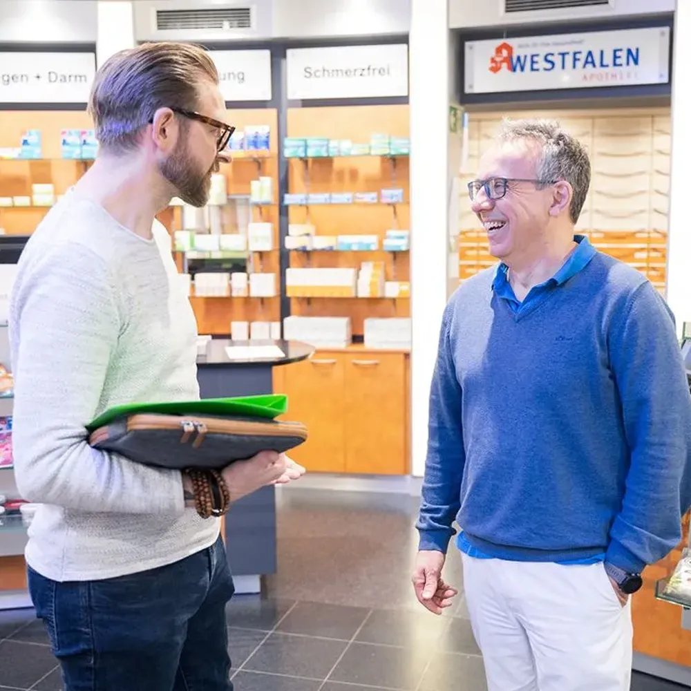 Zwei Männer unterhalten sich freundlich in einer modernen Apotheke vor Regalen mit Medikamenten.
