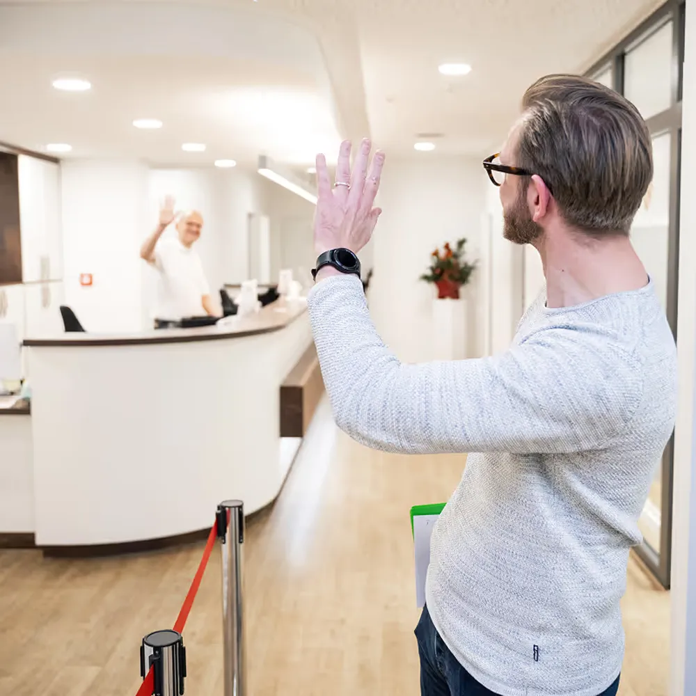 Mann in grauem Pullover winkt einem älteren Mann am Empfang eines lichtdurchfluteten modernen Gebäudes zu.