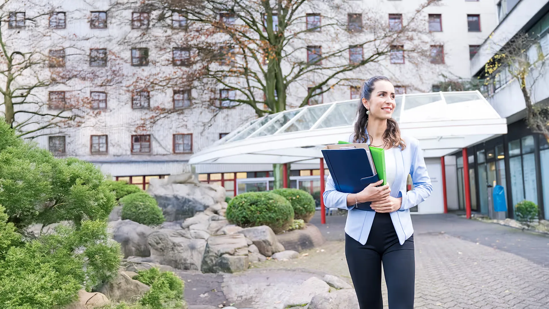 Lächelnde Frau mit Aktenordnern vor einem modernen Gebäude mit Bäumen und grünen Büschen.