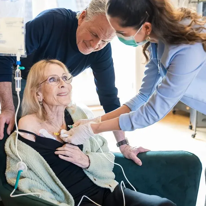 Ärztin mit Maske verbindet eine ältere Patientin, während ein älterer Mann fürsorglich zuschaut.