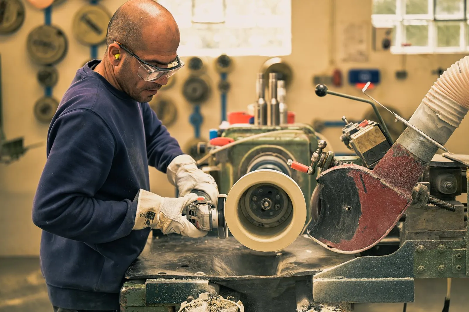 Mann mit Schutzbrille und Gehörschutz arbeitet an einer Schleifmaschine in der Werkstatt.