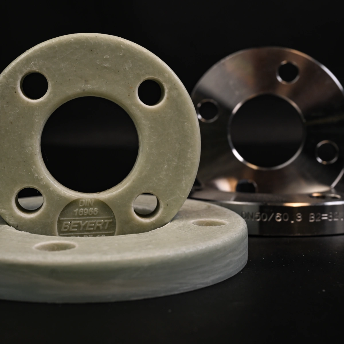 Zwei chemische Rohstoffflansche, einer aus hellem Kunststoff mit sichtbarer Prägung DIN 16966 BEYERT, der andere aus glänzendem Metall mit eingravierter Beschriftung.