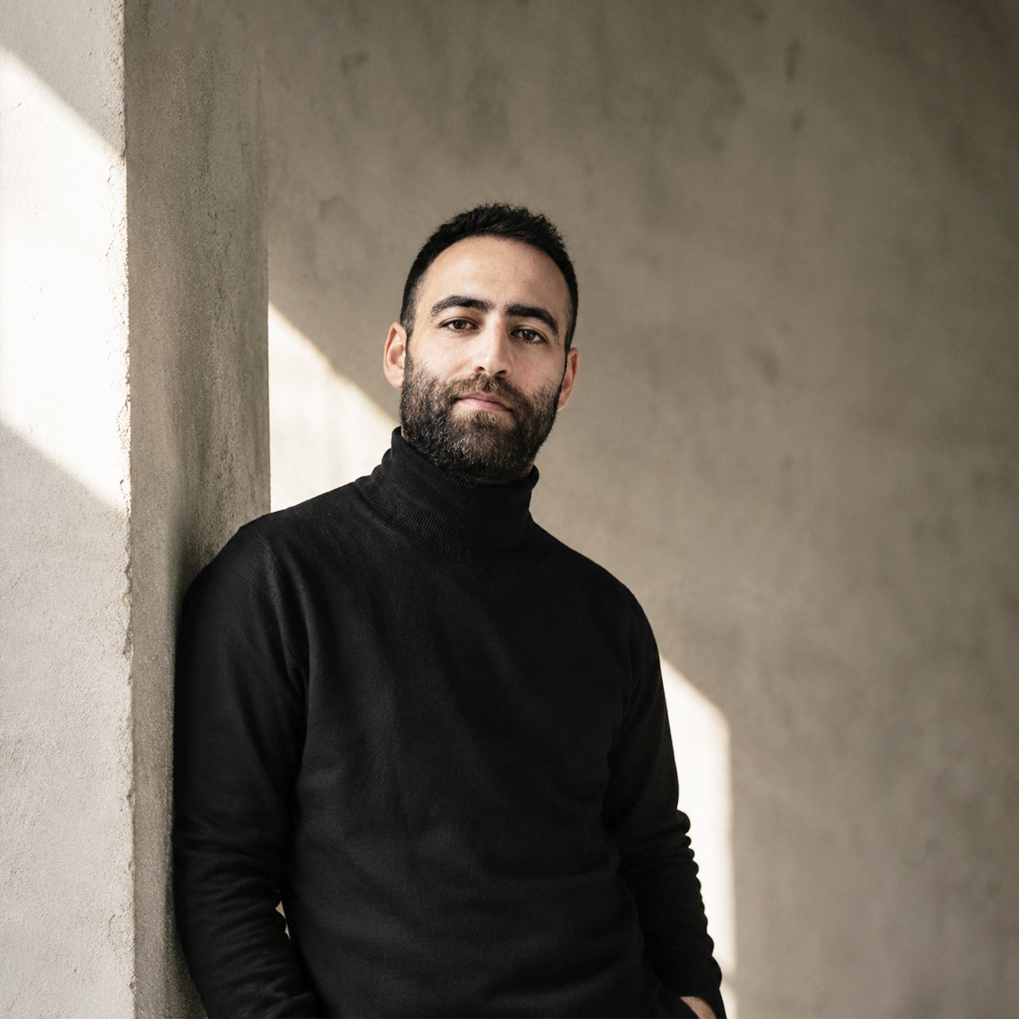 Man with short dark hair and beard wearing a black turtleneck in front of a plain light background.
