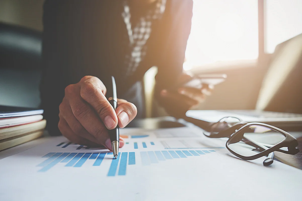 Une personne examine des graphiques financiers tout en tenant un stylo, symbolisant le processus de demande de financement pour une start-up. Des lunettes reposent à côté, suggérant un travail minutieux et réfléchi.