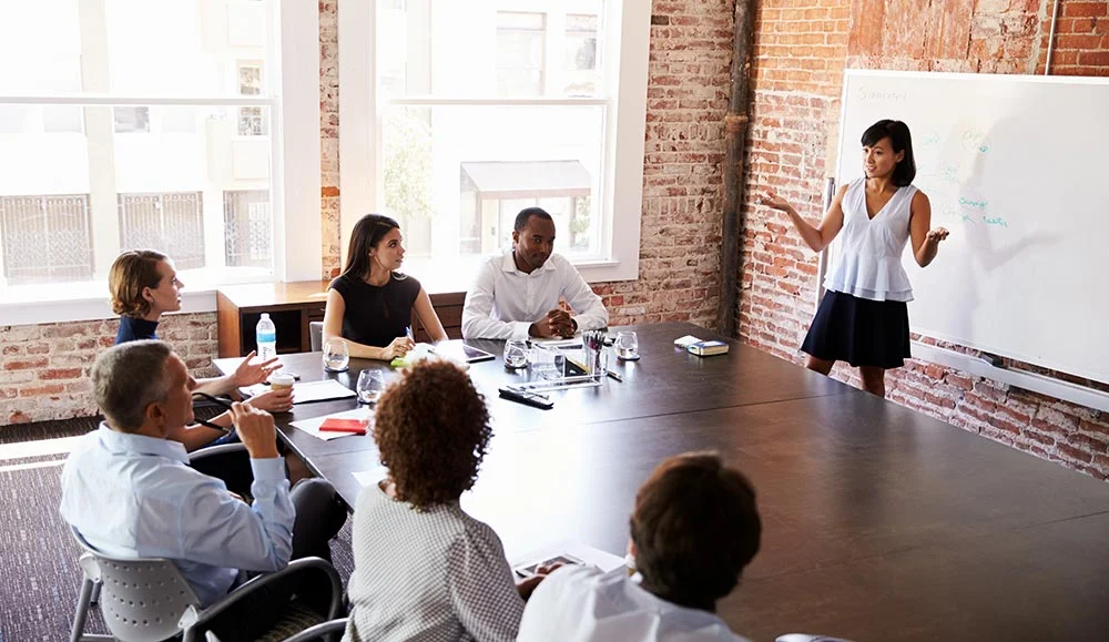 Une personne présente son projet devant un groupe de professionnels, représentant l'étape cruciale de vendre son projet de start-up. La présentation se déroule dans une salle de réunion avec un tableau blanc pour soutenir les idées.