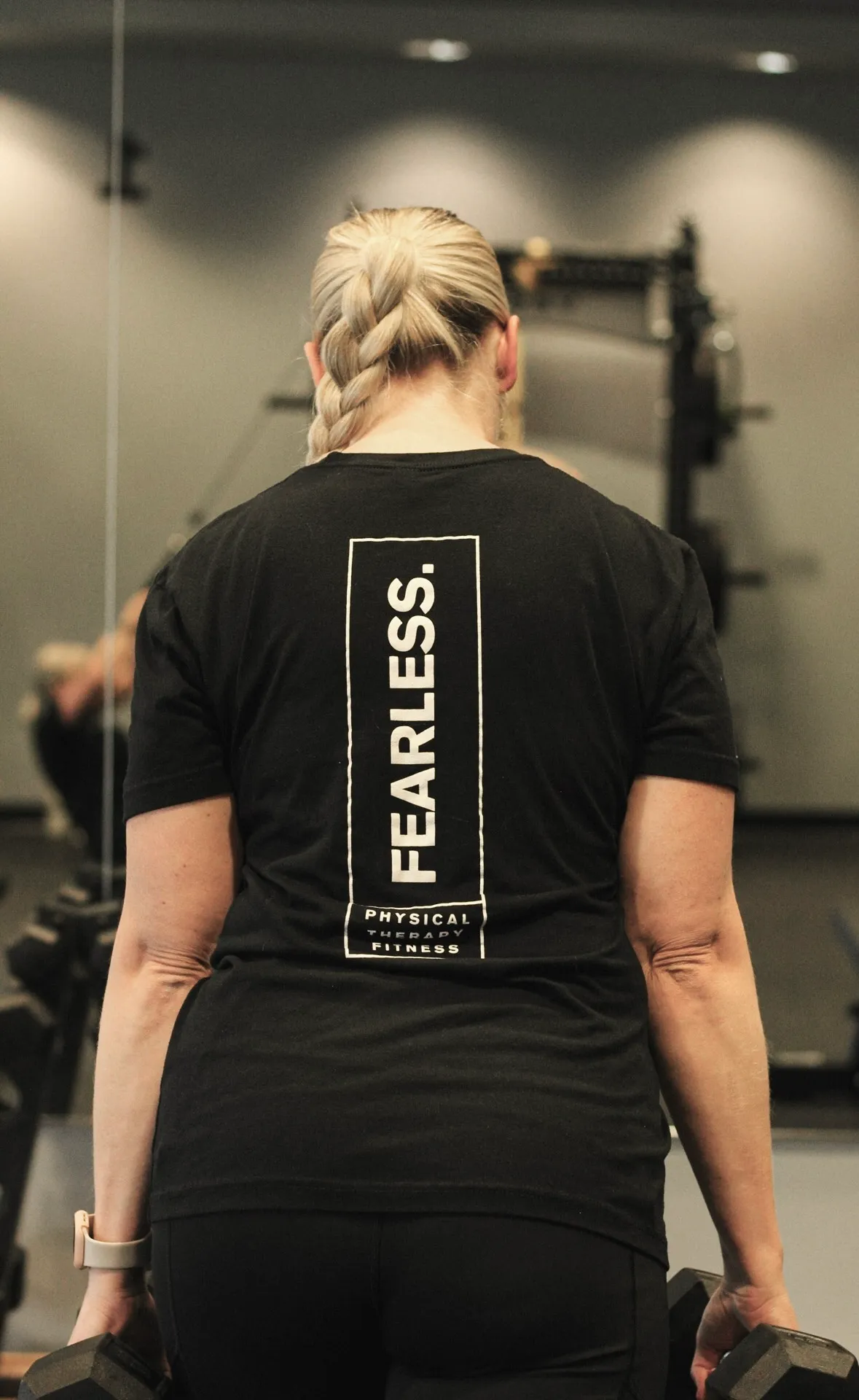 Rear view of a person with a braided ponytail holding dumbbells, wearing a black t-shirt with the text 'FEARLESS. PHYSICAL THERAPY FITNESS' in a gym setting.