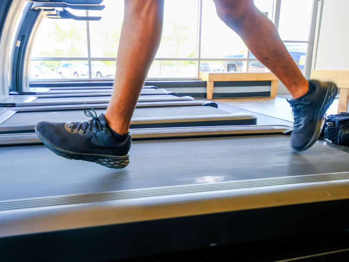 Close-up of a person running on a treadmill in a gym with large windows and parked cars outside.