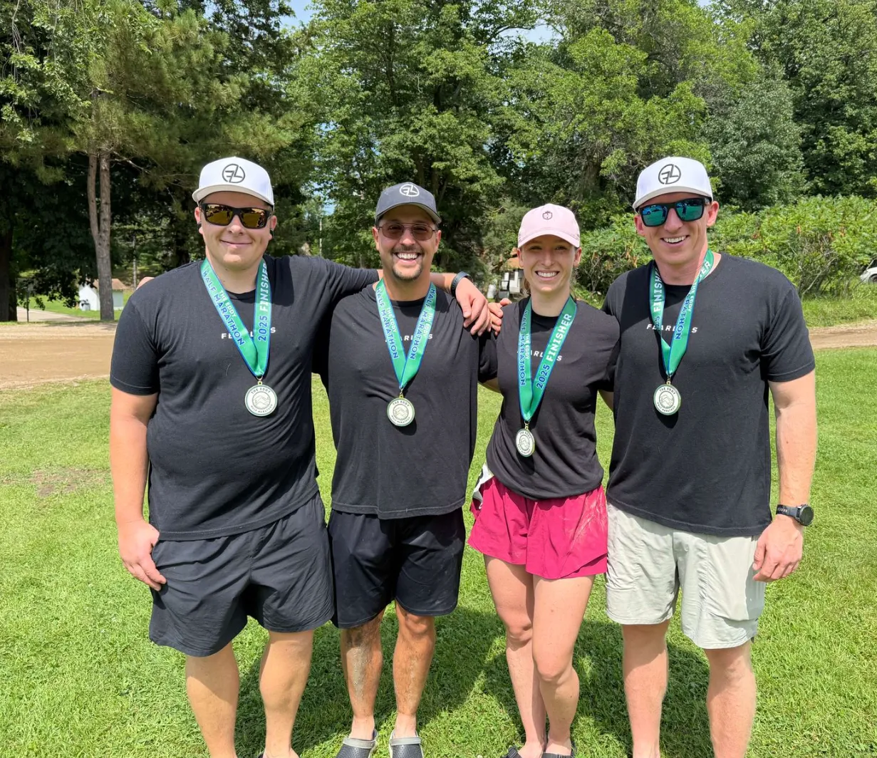 Four people standing outdoors on grass wearing finisher medals from the 2025 half marathon, dressed in black athletic shirts and hats, smiling at the camera.