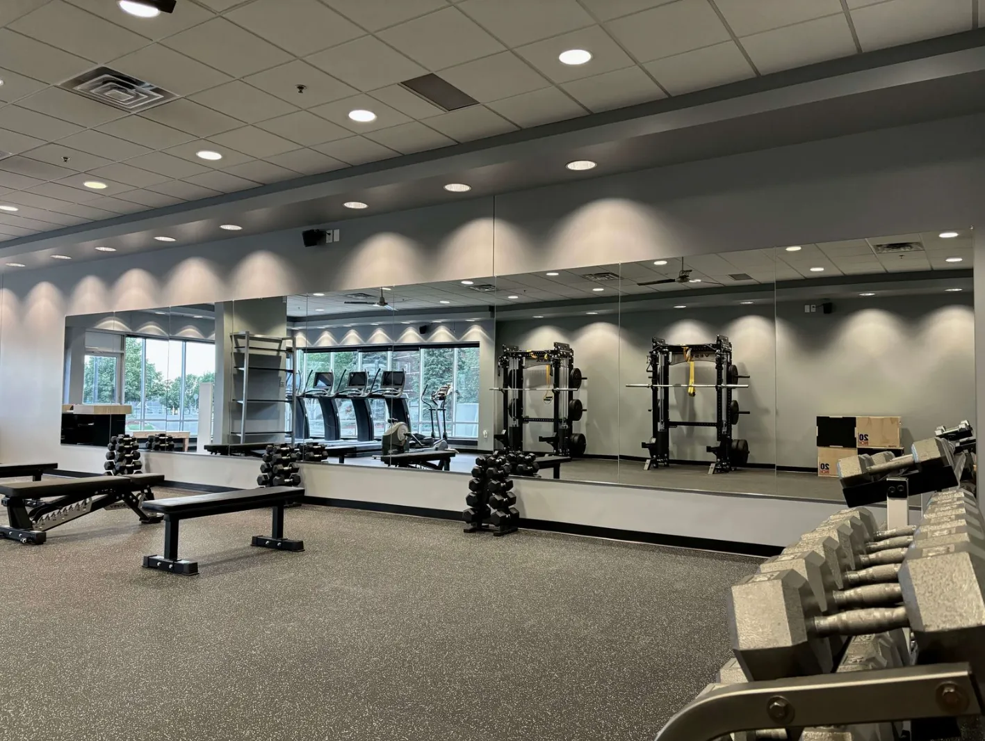 Modern gym interior with benches, dumbbells, treadmills, and squat racks reflected in large wall mirrors.