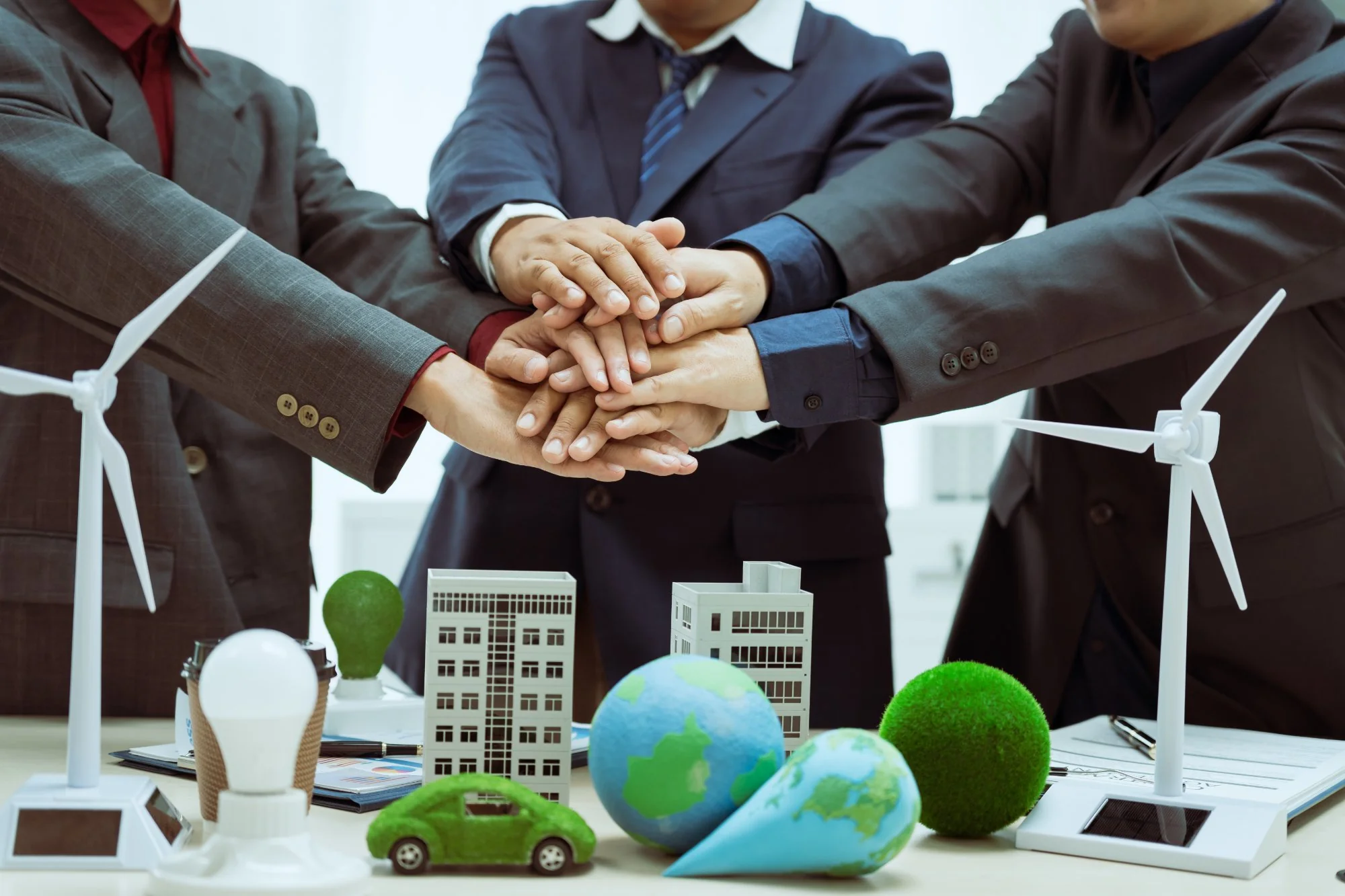 Three businesspeople joining hands over a table with sustainability models including wind turbines and a globe, representing environmental compliance partnership