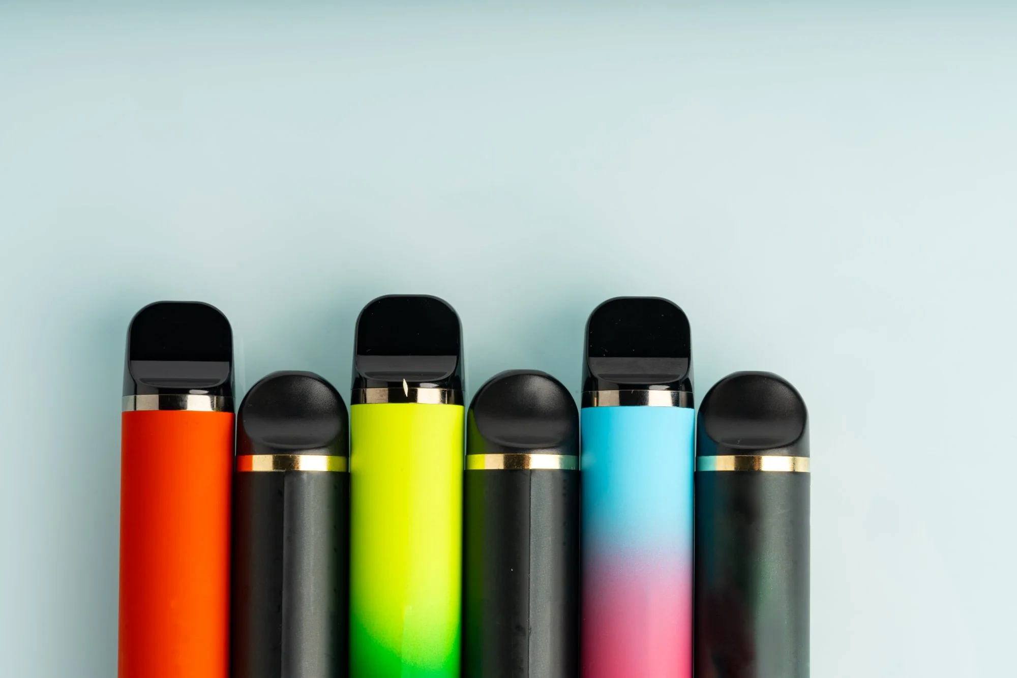 Row of colourful disposable electronic cigarettes and vape devices on a light blue background representing Category 15 WEEE products