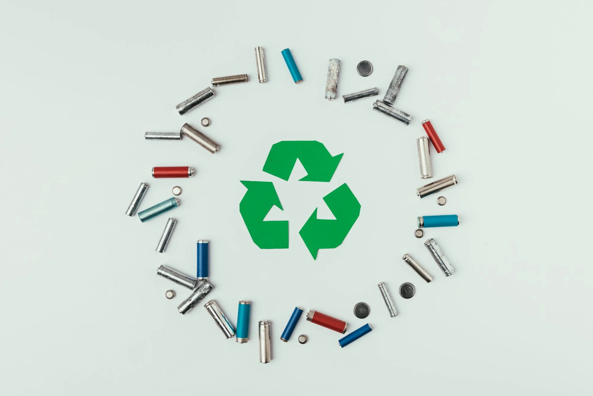 Top view of various types of batteries including AA, AAA and button cells arranged around a green recycling symbol