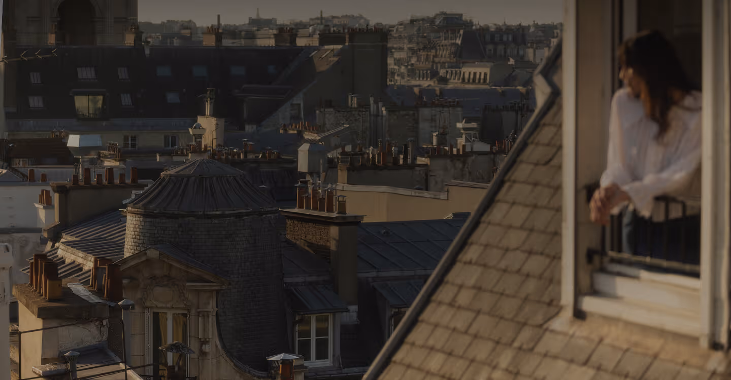 Une femme appuyée sur le rebord d'une fenêtre regarde les toits en ardoise d'un quartier urbain dense au coucher du soleil. - Hôtel Madison