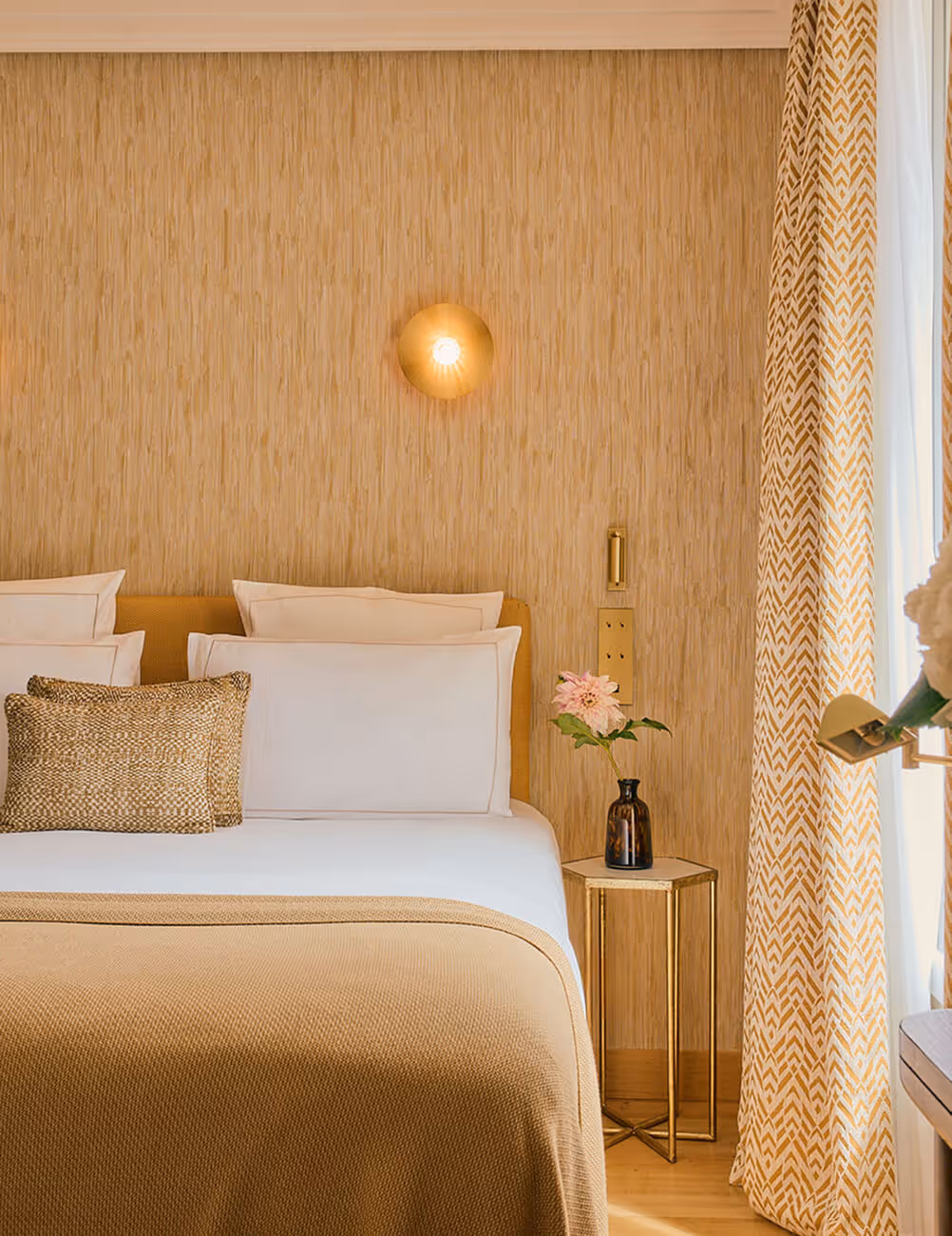 Chambre élégante avec lit double beige, coussins décoratifs, table de chevet avec vase de fleurs, et rideaux à motifs jaunes. - Hôtel Madison