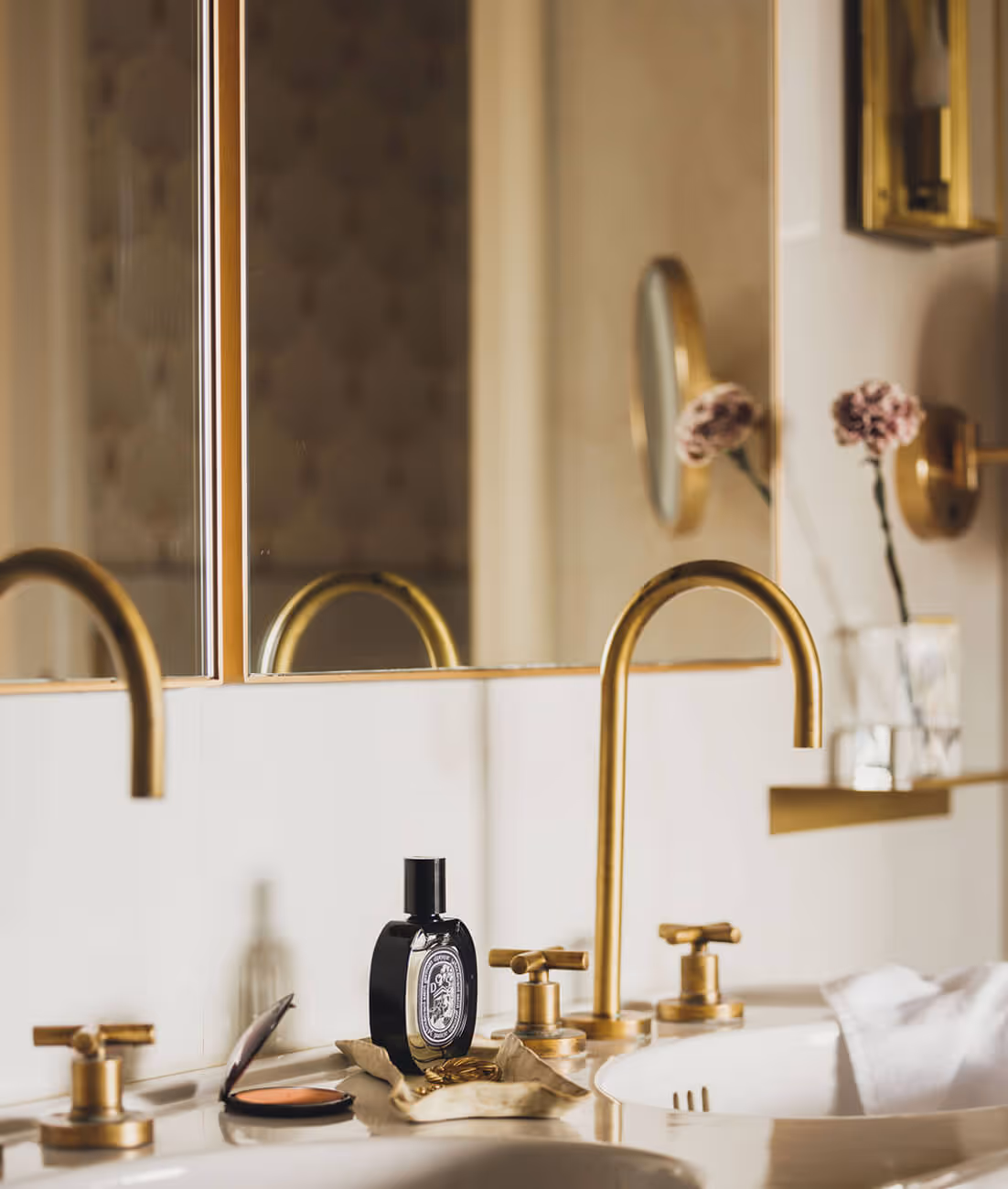 Lavabo double avec robinets dorés, flacon de parfum noir, miroir et fleur rose en arrière-plan. - Hôtel Madison