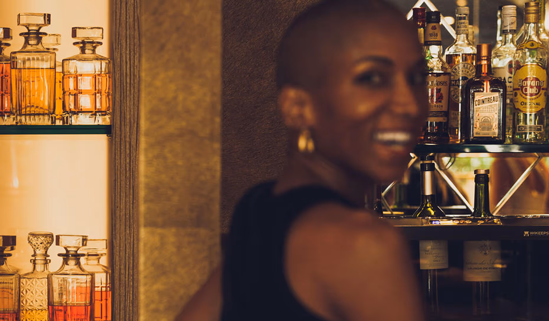 Personne souriante avec boucle d'oreille dorée devant un bar avec des bouteilles d'alcool et des carafes en verre. - Hôtel Madison
