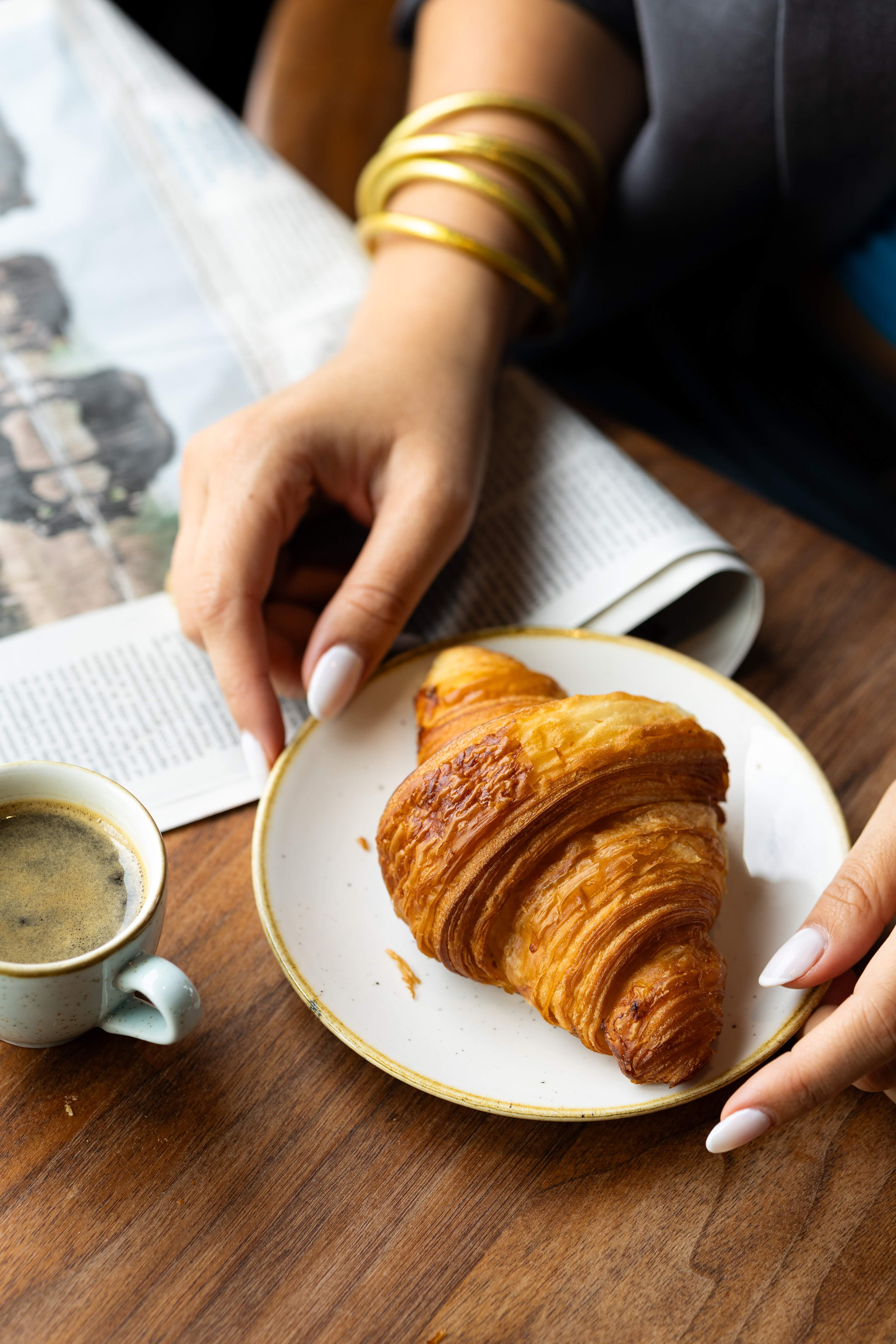 Hôtel Madison - Une personne avec des bracelets dorés tient une assiette blanche avec un croissant doré à côté d'une tasse de café sur une table en bois.