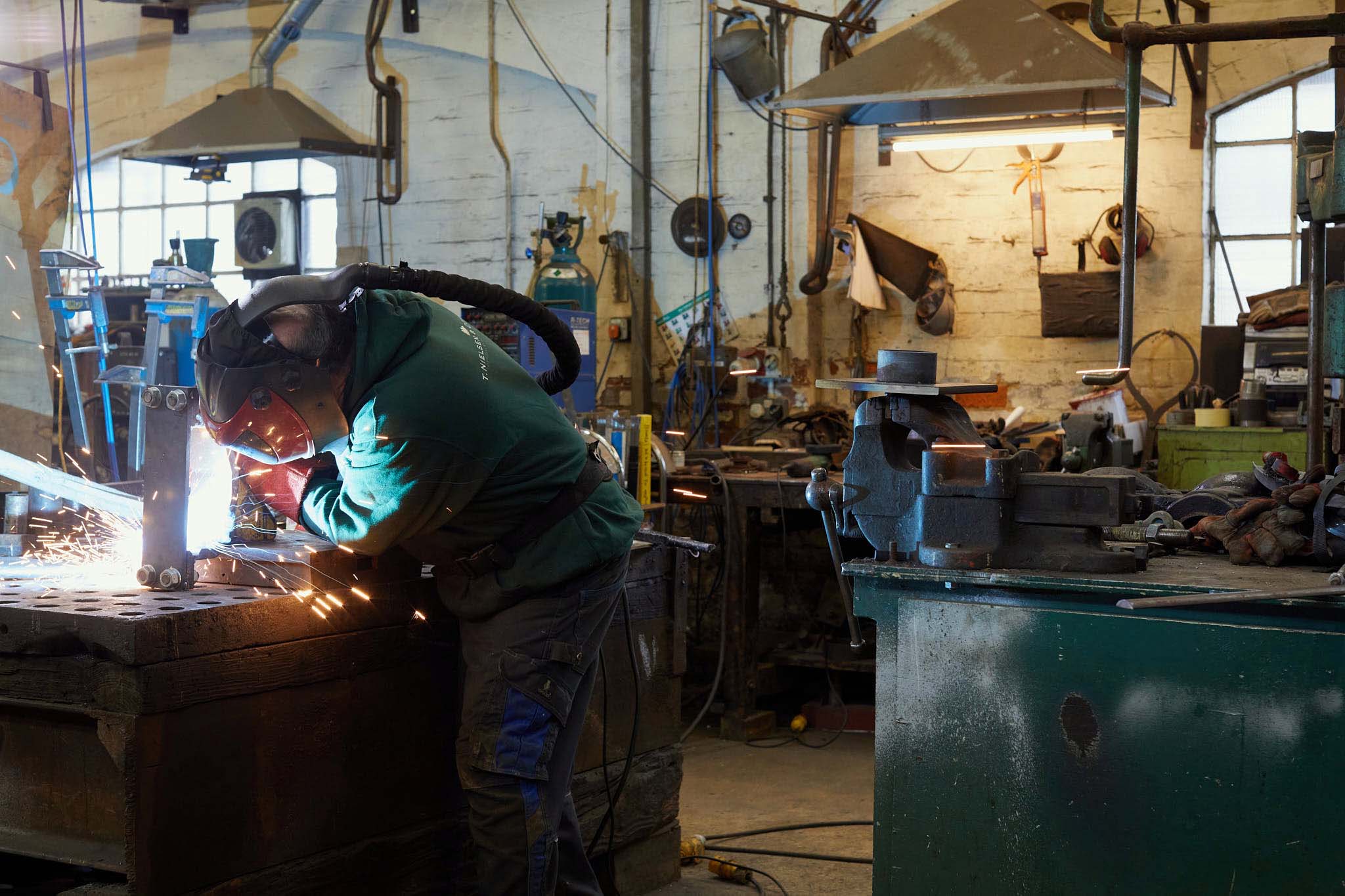 T Nielsen and Co Man in protective mask and helmet welding at table sparks surrounded by factory machinery
