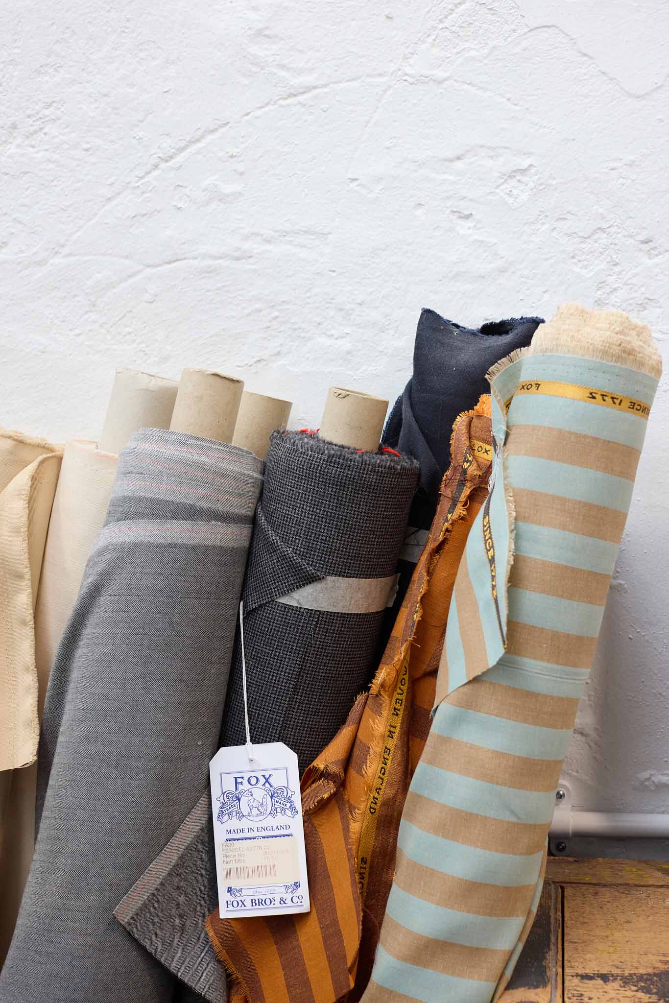 Fox Brothers rolls of cloth leaning against white wall the cloth is grey charcoal orange and stiped orange there is a large white label in the centre