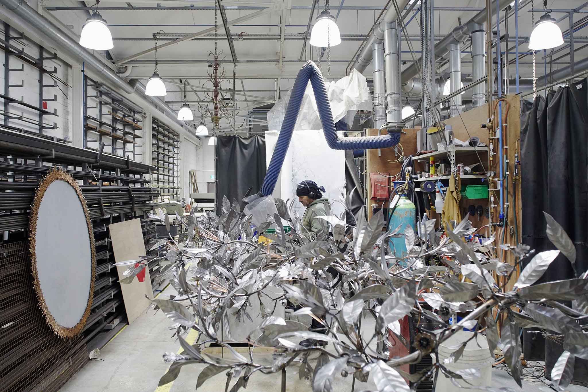 Cox London inside workshop with silver metalwork leaf sculpture in foreground employee behind and large gold sunburst mirror on the wall on the left of the picture