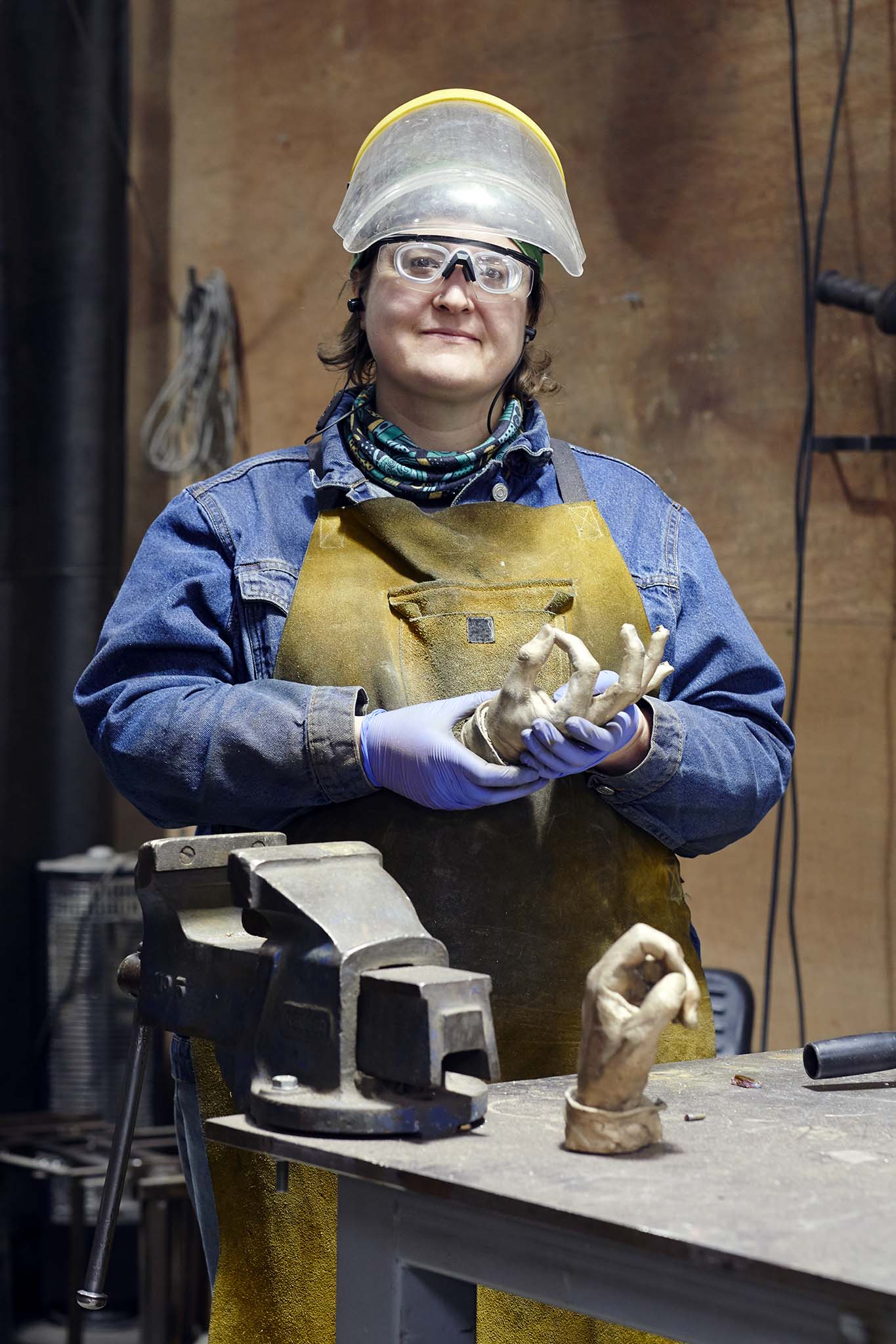 Cox London woman in blue overalls and protective eyewear standing behind work table with sculpture of golden hand on table 