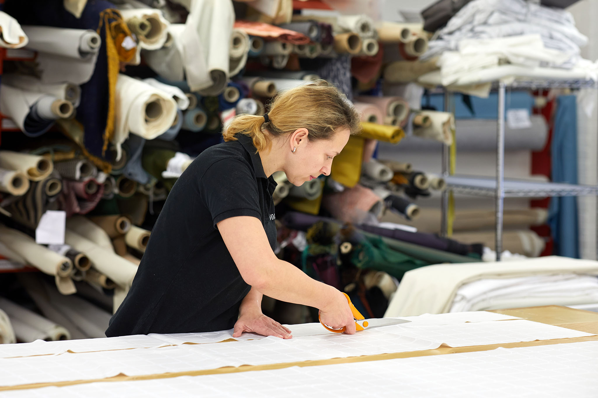 Savoir Beds woman cutting fabric at table with shelves full of rolls of fabric behind her