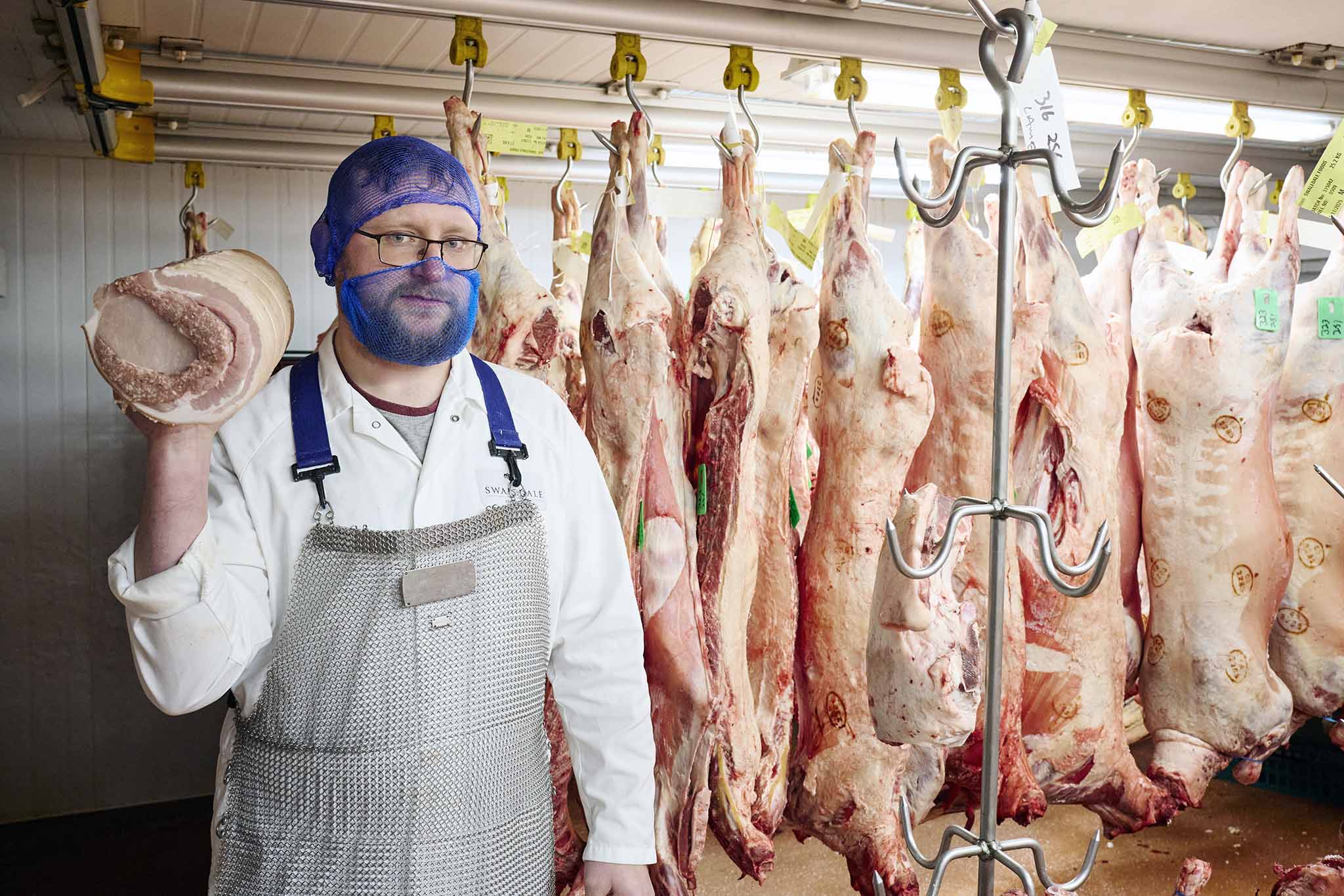 Swaledale Butchers butcher to the left of the picture holding piece of meat with large carcasses hanging from hooks in the background 