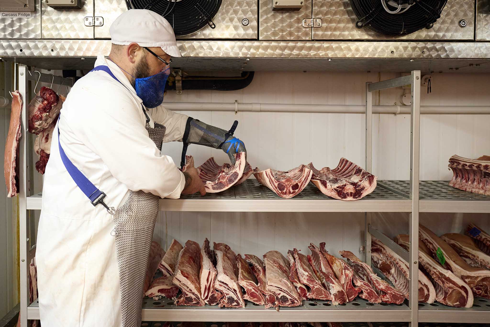 Swaledale Butchers butcher on left of picture in apron and white hat blue mask shelves of cuts of meat