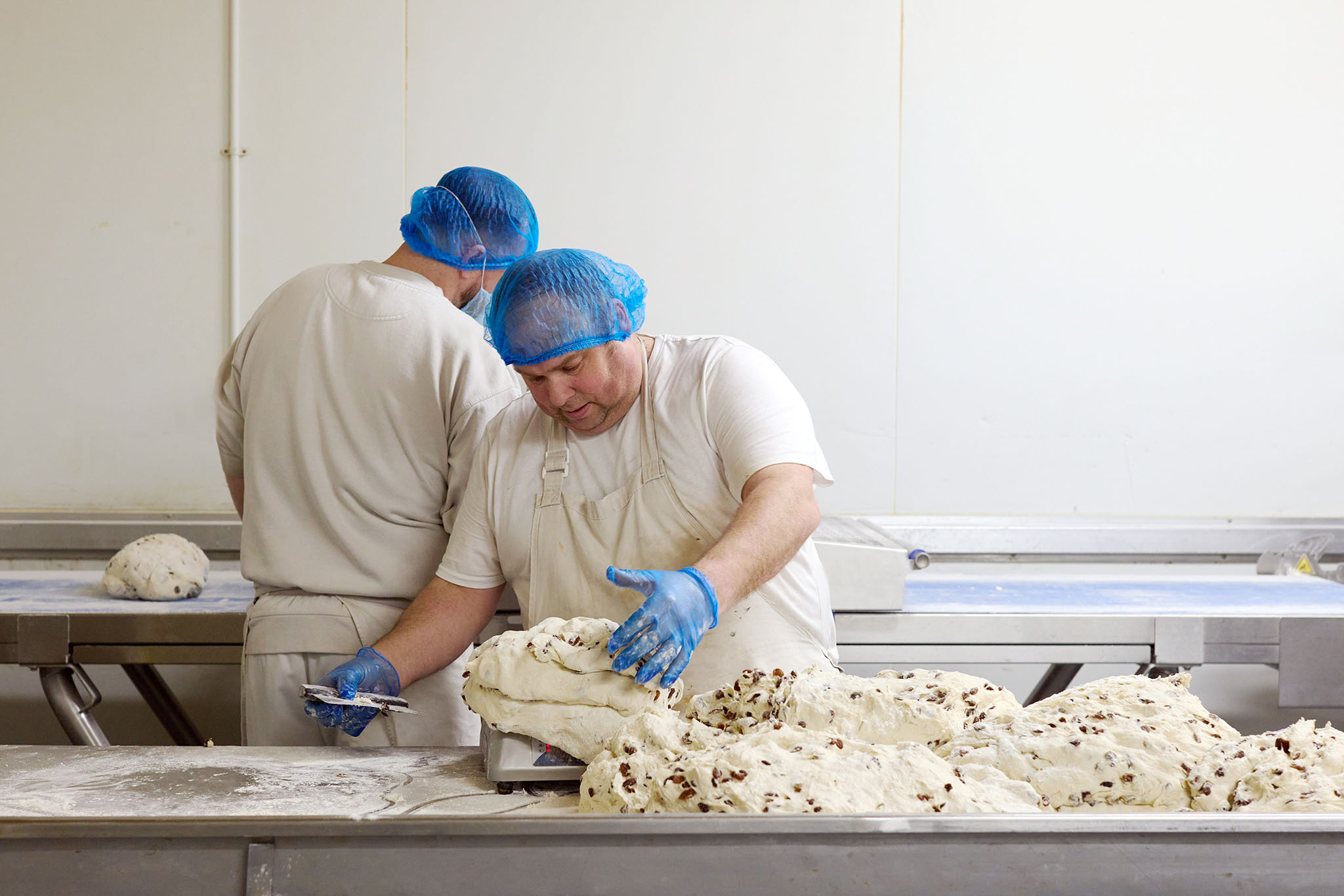 Janes Pantry two men in white clothing and blue hair nets and aprons measuring out dough full of sultanas or raisins dough is on workbench