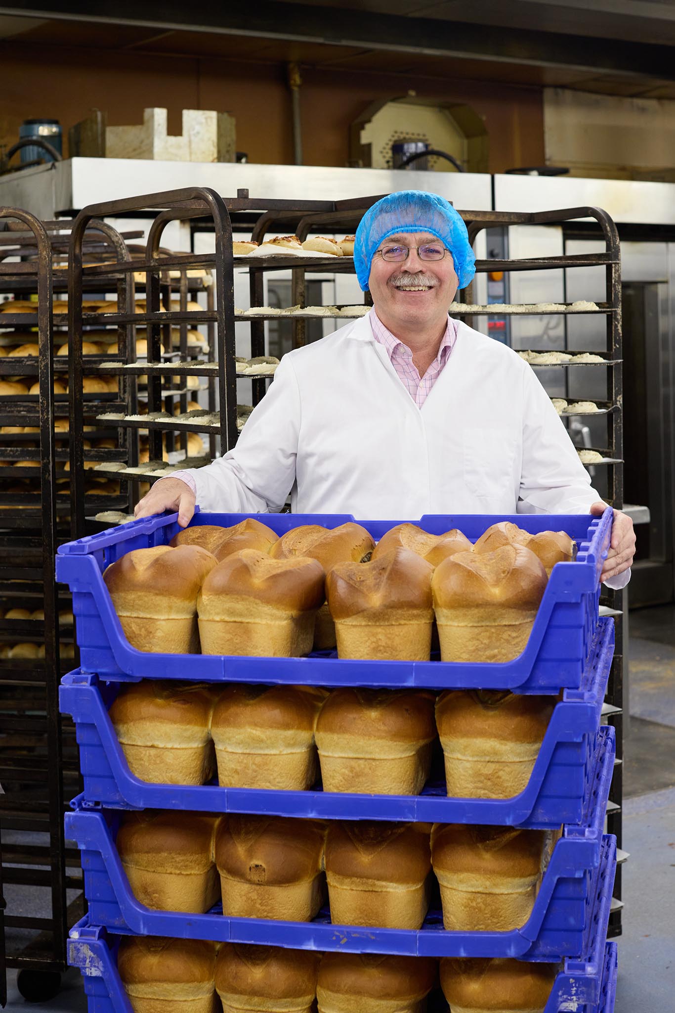 Janes Pantry smiling man in blue hair net and white coat standing behind stacked blue trays of loaves of white bread