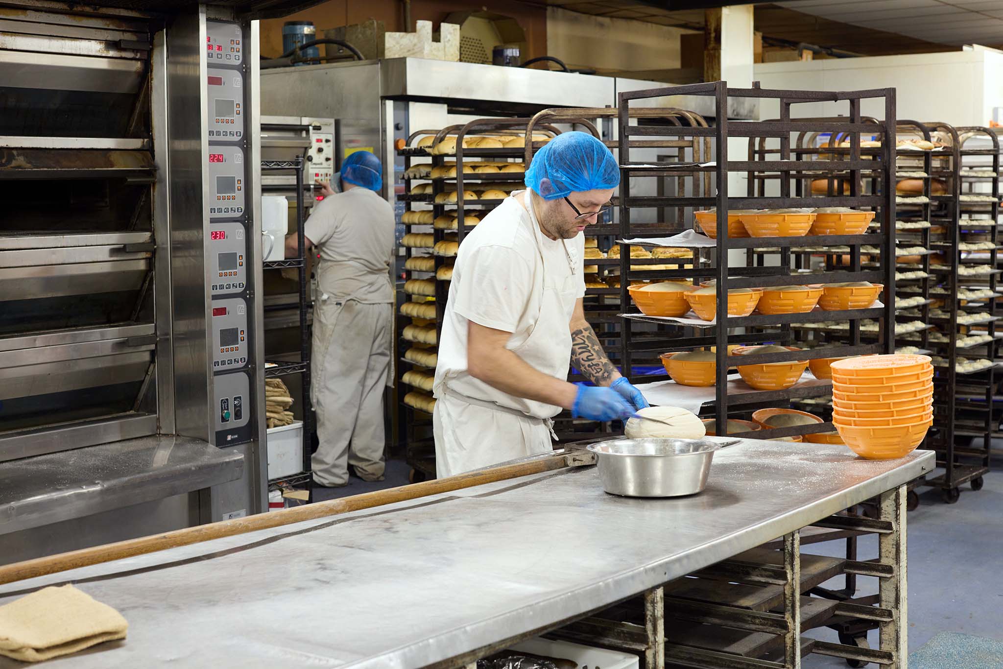 Janes Pantry employee baker finalising uncooked loaf of bread trolley behind him full of orange coloured bread baskets