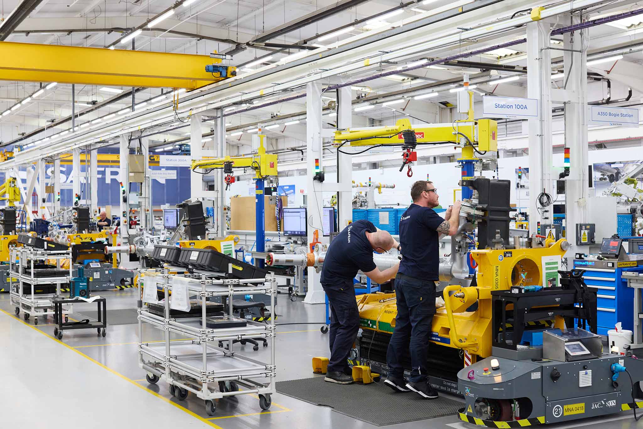 Safran Landing Systems inside factory full of large machines yellow metal trollies full of tools two men in blue uniform working at yellow machine