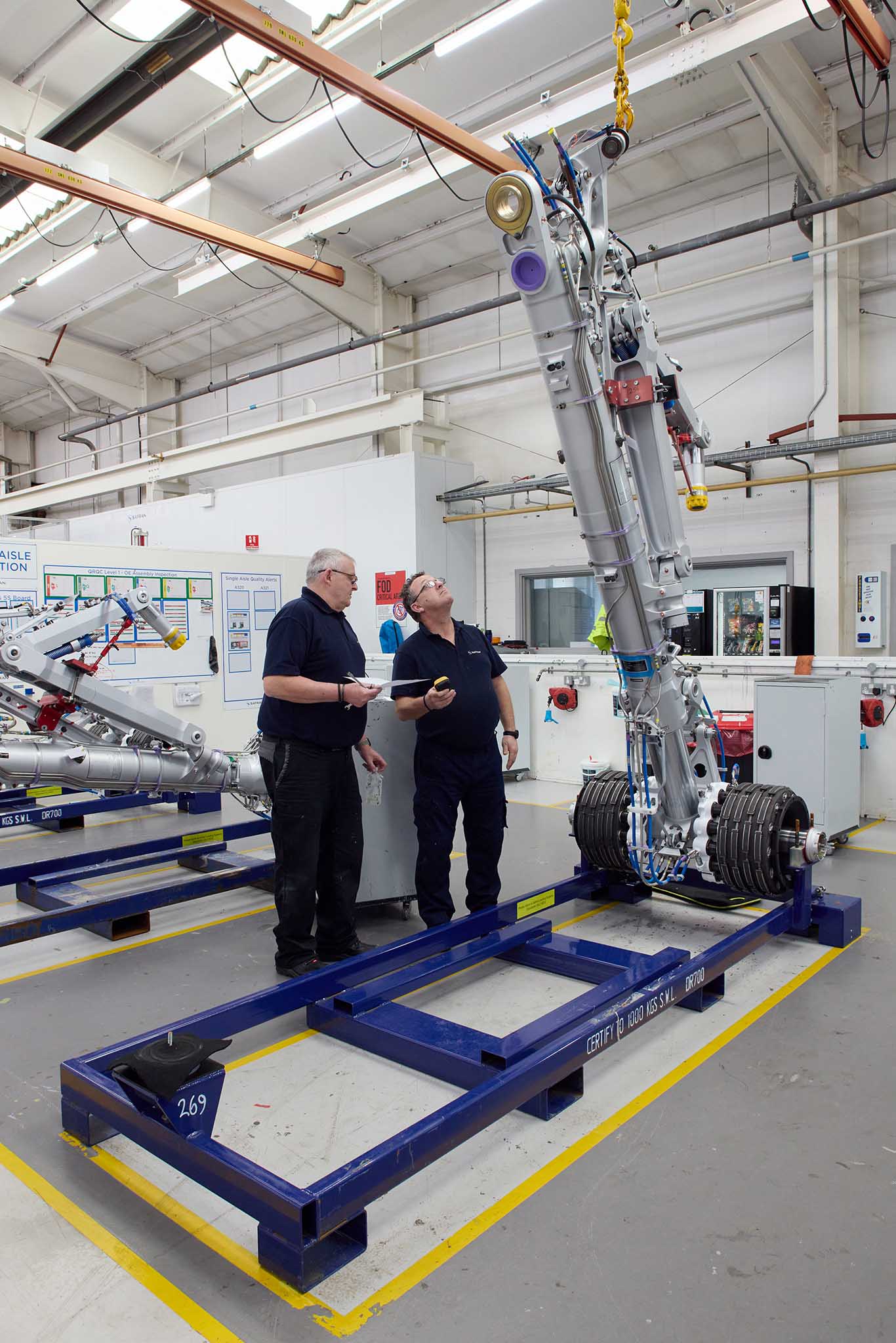 Safran Landing Systems inside factory tall ceilings two employees engineers in uniform working at large metal machinery 