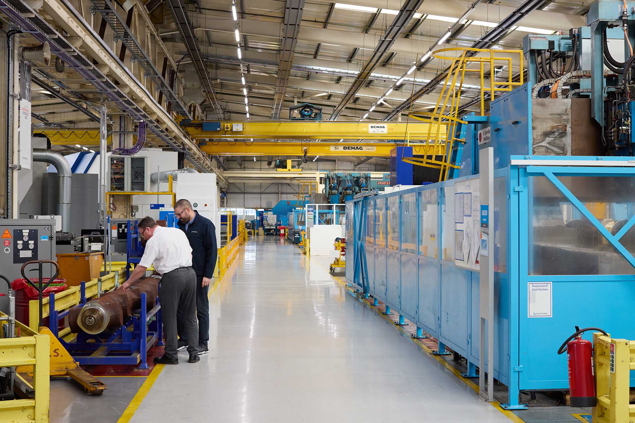 Safran Landing Systems inside of large factory with clear floors and high ceiling and strip lighting there are yellow steel  metal machines and blue machines