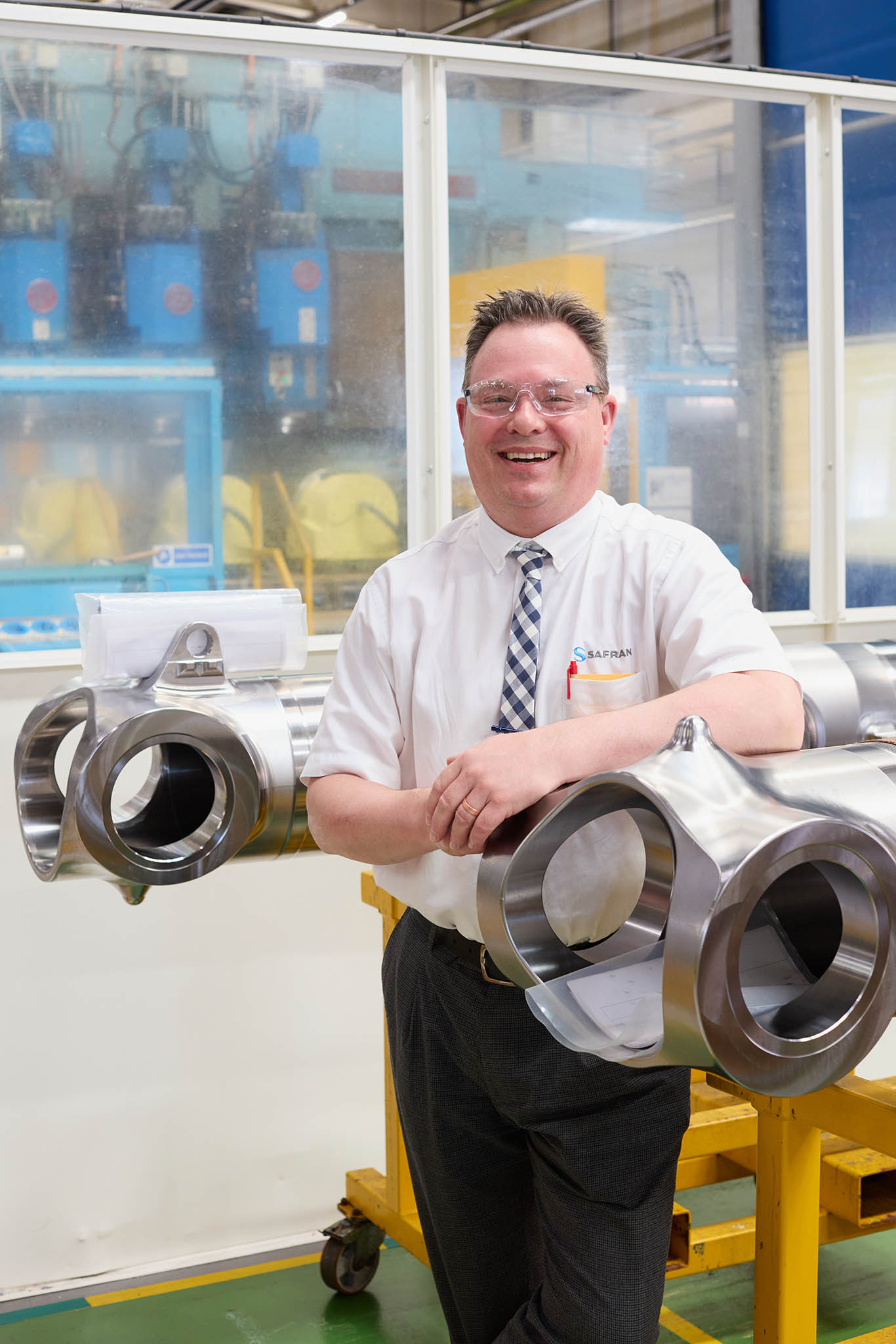 Safran Landing Systems engineer employee in in white shirt and tie standing behind metal aircraft componentry 