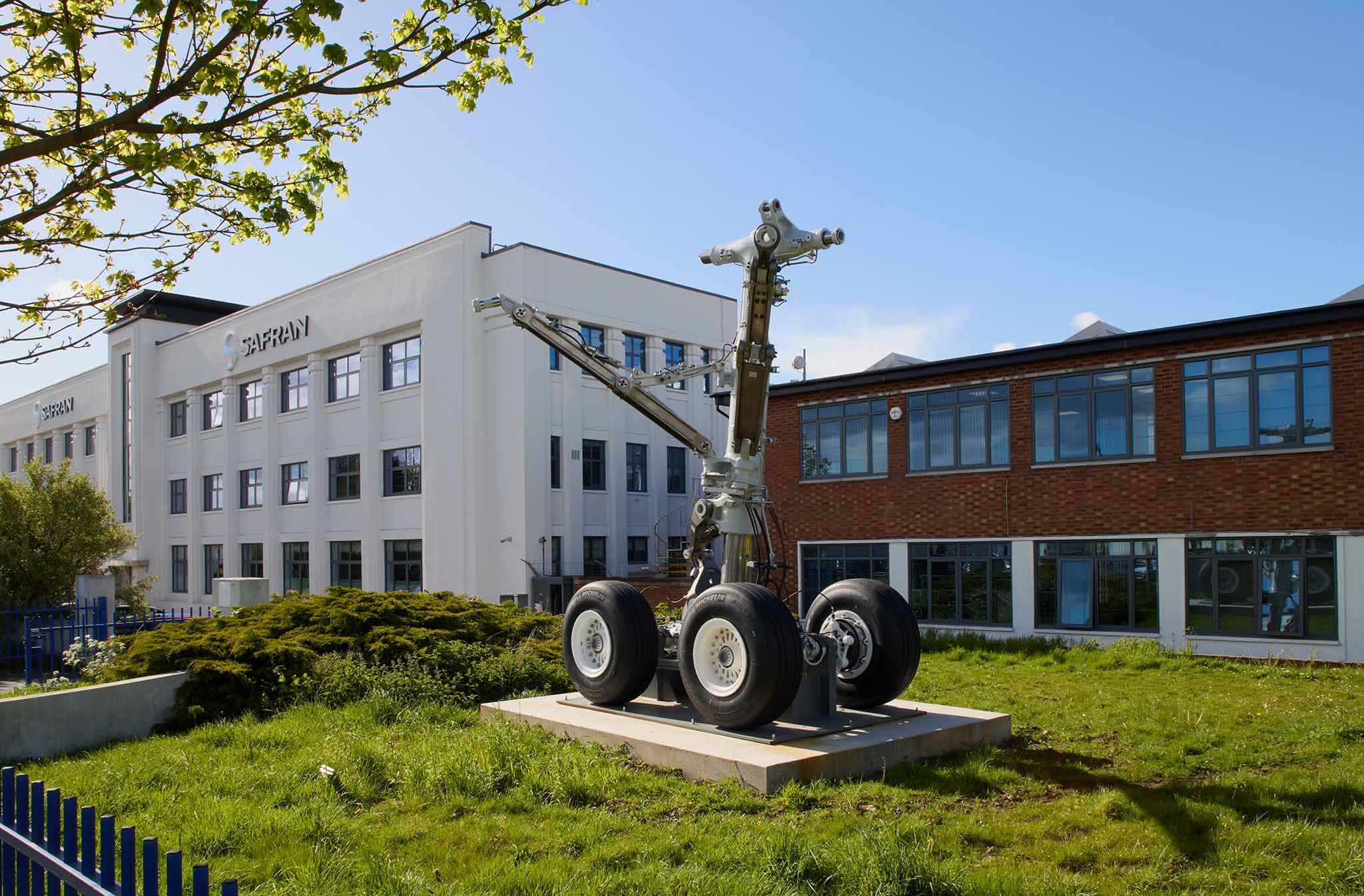 Safran Landing Systems outside factory buildings grass in foreground with aircraft landing gear on platform on lawn there is white three storey building to the left and a red brick building to the right