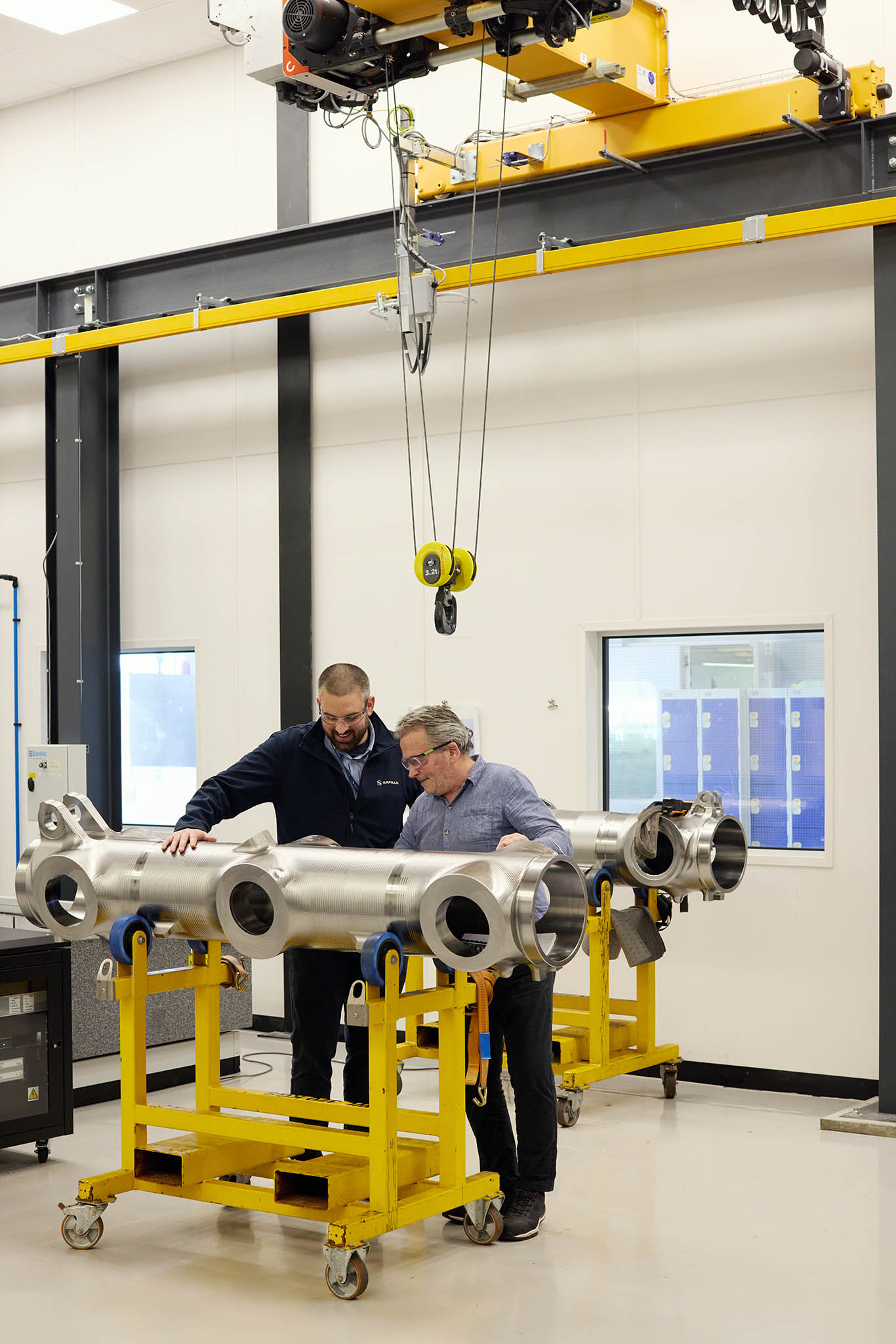 Safran Landing Systems two engineers employees in protective glasses standing behind yellow metal trolly working with tool on large metal componentry pipes aircraft
