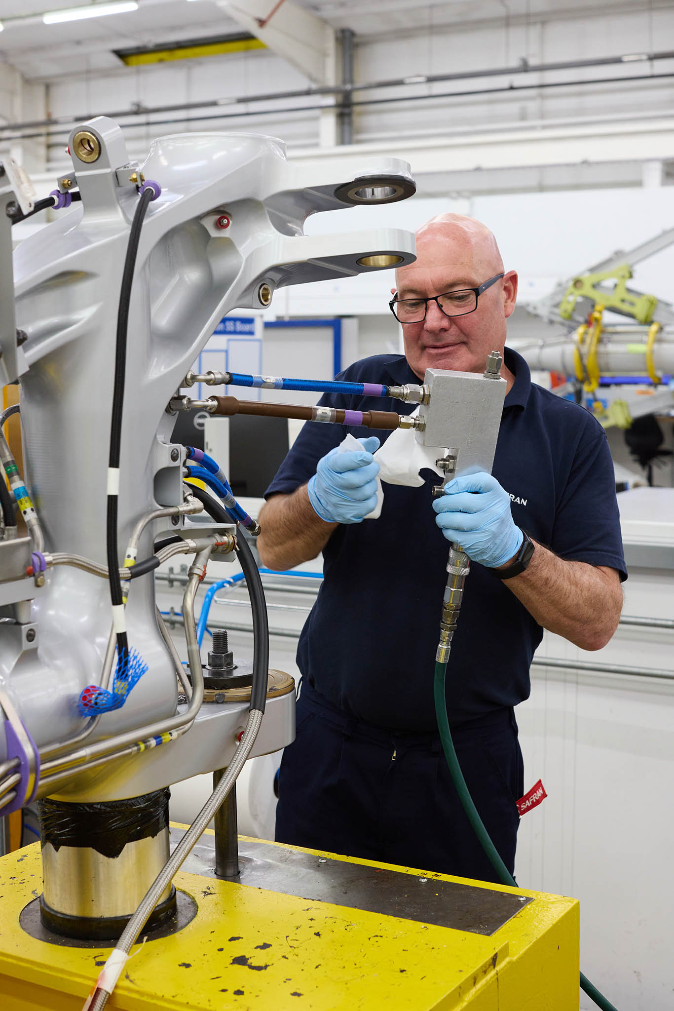 Safran Landing Systems engineer employee in protective glasses standing behind yellow machine working with tool on metal componentry pipes aircraft