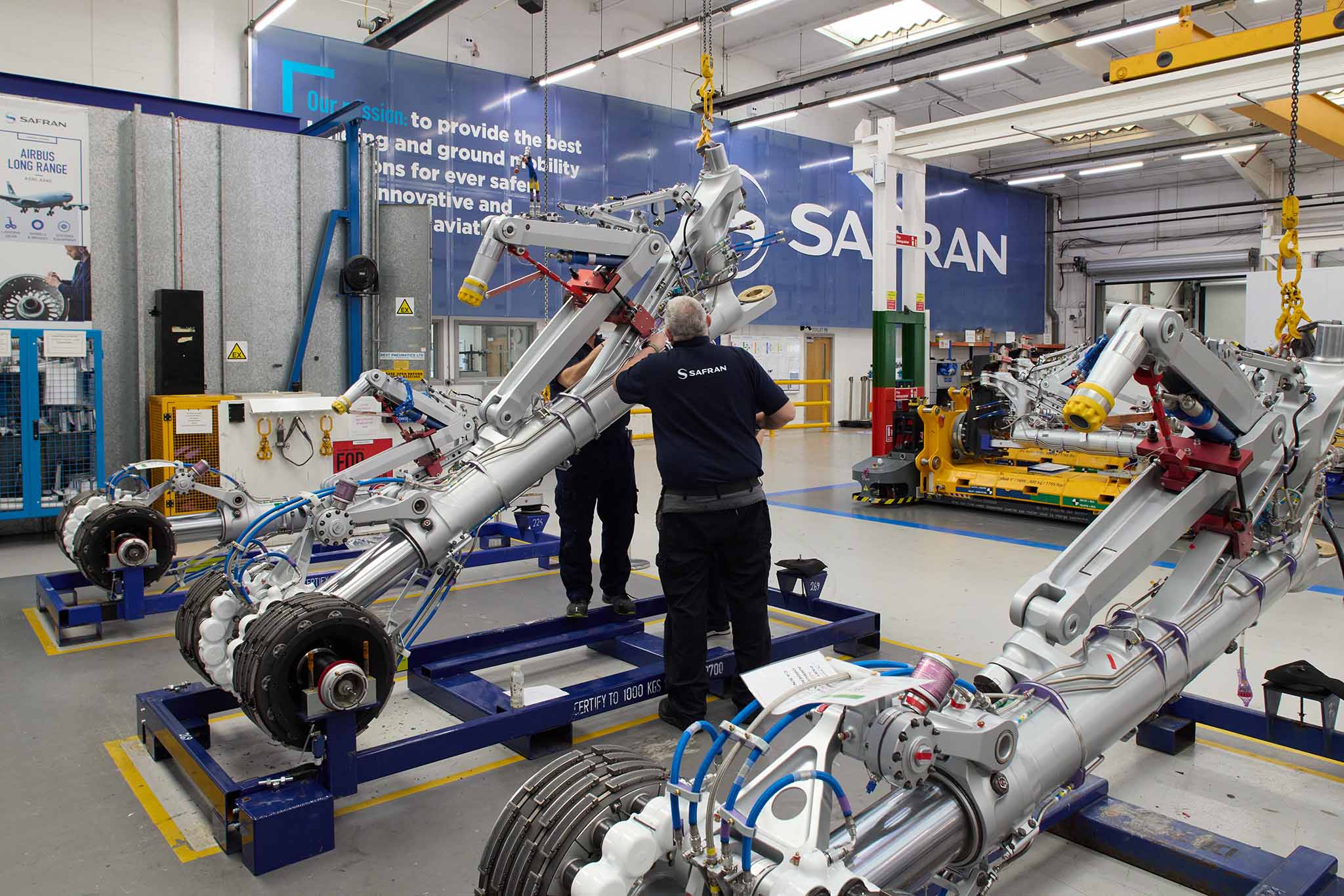 Safran Landing Systems inside of large factory with clear floors and high ceiling and strip lighting there are yellow steel metal machines and blue machines